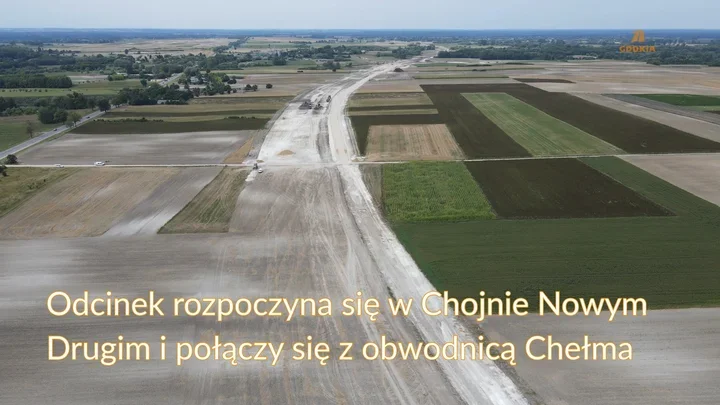 Widok z lotu ptaka na budowę drogi przez pola uprawne, z nanieszonym logotypem GDDKIA.