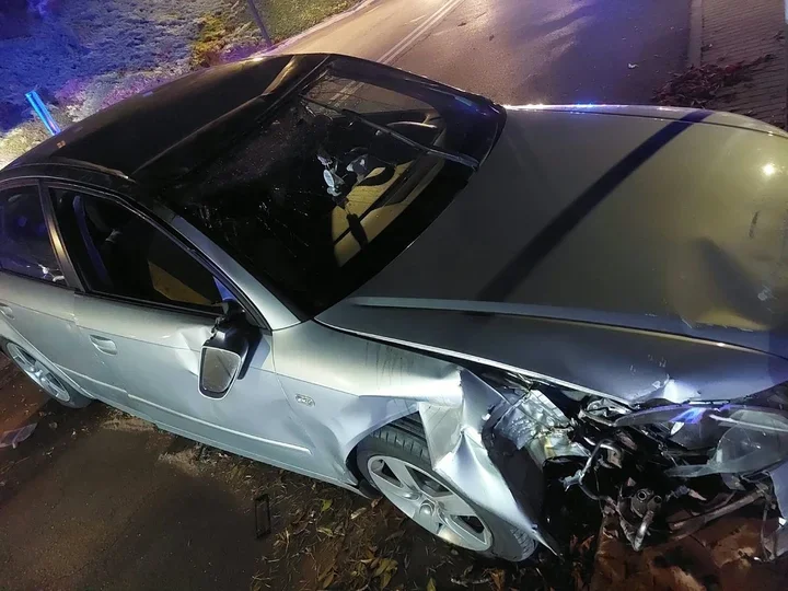 Uszkodzony srebrny samochód osobowy stoi nocą przy drodze po zderzeniu, z widocznym zniszczeniem przedniej części pojazdu.