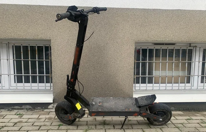 Elektryczna hulajnoga stoi na chodniku przed szarą elewacją budynku z oknami zabezpieczonymi kratami.