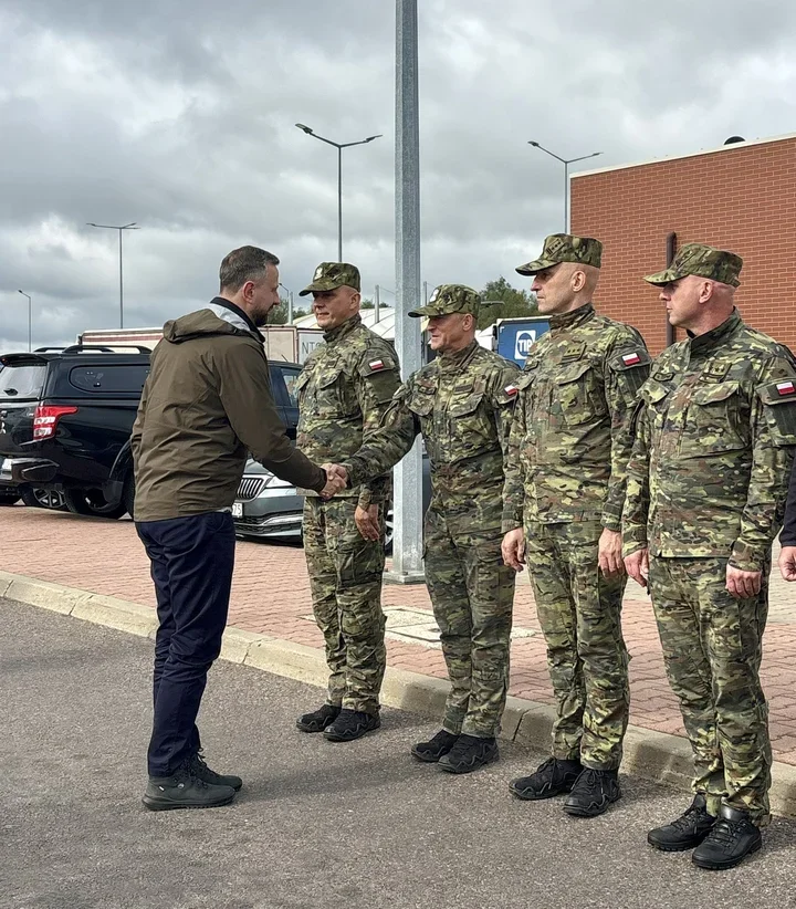 Mężczyzna w brązowej kurtce wita się uściskiem dłoni z żołnierzem w kamuflażu, stojącym w szeregu z trzema innymi żołnierzami, na parkingu przed ceglanym budynk