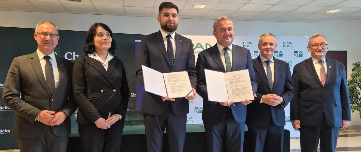 Sześć osób w garniturach stoi podczas uroczystości podpisania dokumentu; dwaj mężczyźni w centrum trzymają otwarte umowy.