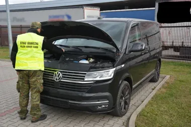 Funkcjonariusz Straży Granicznej sprawdza silnik czarnego volkswagena z otwartą maską na brukowanym placu przy ogrodzeniu.