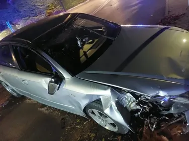 Uszkodzony srebrny samochód osobowy stoi nocą przy drodze po zderzeniu, z widocznym zniszczeniem przedniej części pojazdu.
