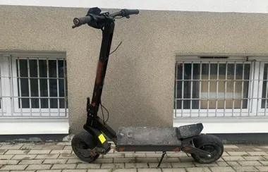 Elektryczna hulajnoga stoi na chodniku przed szarą elewacją budynku z oknami zabezpieczonymi kratami.
