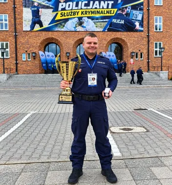 Policjant w mundurze stoi przed budynkiem z banerem „Policjant Służby Kryminalnej”, trzymając duży złoty puchar i medal.