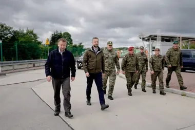 Dwaj mężczyźni w ubraniach cywilnych idą przed grupą żołnierzy w mundurach po terenie infrastrukturalnym pod szarym niebem.
